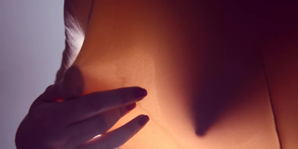 Eine Nahaufnahme zeigt eine Hand, die ein Stück transparenten, orangefarbenen Stoff sanft berührt. Das Licht von hinten lässt die feine Struktur des Stoffes erkennen und wirft den Schatten der Hand deutlich auf die Oberfläche.
