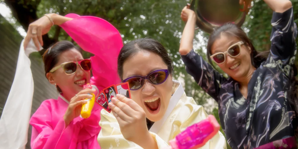 Drei Personen in bunten Hanbok, traditionelle koreanische Kleidung mit weiten Ärmeln und leuchtenden Farben, und Sonnenbrillen posieren im Freien, eine Person zeigt Spielkarten.