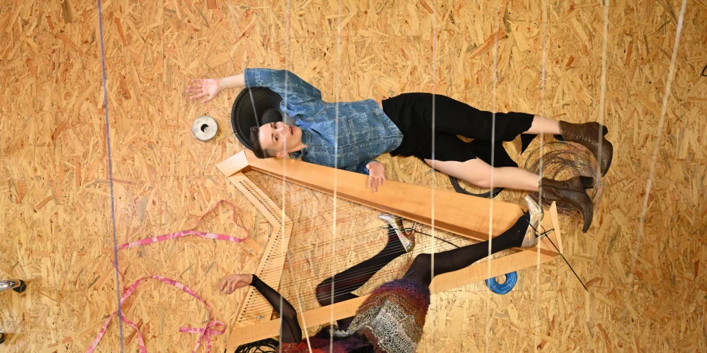 Two people lie on a wooden floor between cables and a large wooden string structure. One person wears a blue denim top, the other a multicolored knitted top with sheer sleeves.