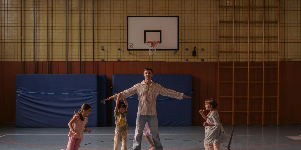 Eine Person steht mit ausgebreiteten Armen in der Mitte einer Turnhalle, während mehrere Kinder um sie herum spielen und laufen. Die Szene ist eingebettet in eine alltägliche Sporthallenumgebung mit blauen Matten, einer Sprossenwand und einem Basketballkorb im Hintergrund.. 