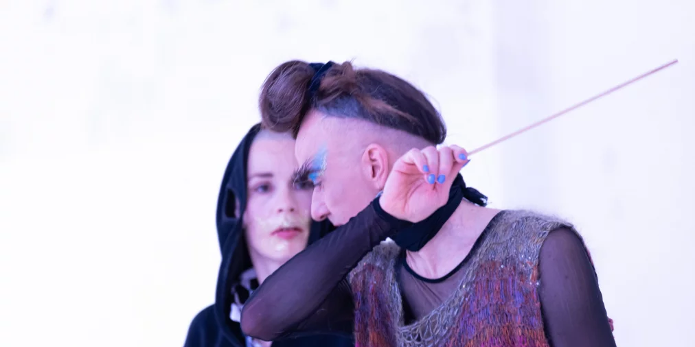 Zwei Performer*innen in einer hell ausgeleuchteten Szene. Eine Person mit hochgestecktem Haar, blauem Lidschatten und transparentem, buntem Oberteil hält einen dünnen Stab und hebt den Arm. Dahinter steht eine Person mit Kapuzenpullover.