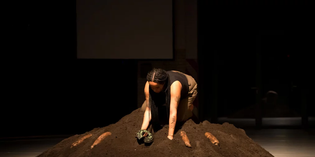 A performer kneels on a large mound of earth, digging into the soil with their hands. The light highlights their posture while the surrounding space remains dark. Behind them, the outline of a projection screen is visible.