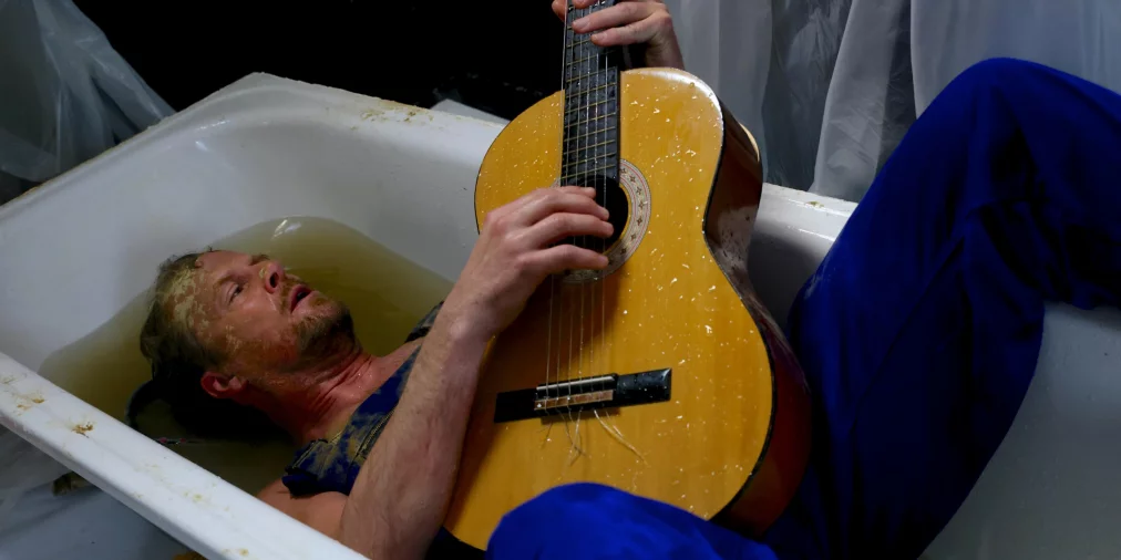 Eine Person liegt in einer mit Wasser gefüllten Badewanne und spielt eine Gitarre, während Arbeitskleidung und Requisiten die Szene umgeben.