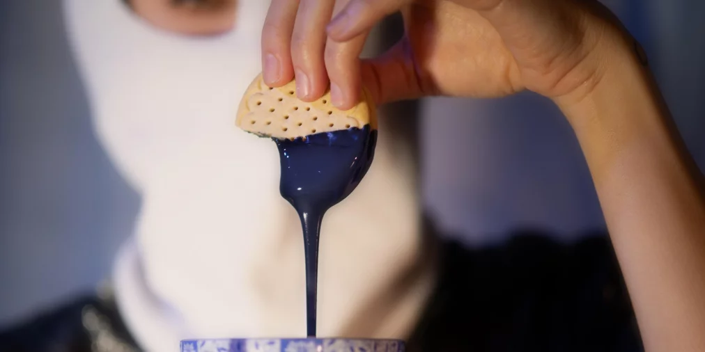 Eine Person hält eine blau-weiß gemusterte Teetasse auf einer Untertasse. Ein Keks wird in eine dickflüssige, dunkelblaue Substanz getaucht, die langsam in die Tasse zurückläuft. Der Oberkörper der Person ist unscharf im Hintergrund zu sehen.