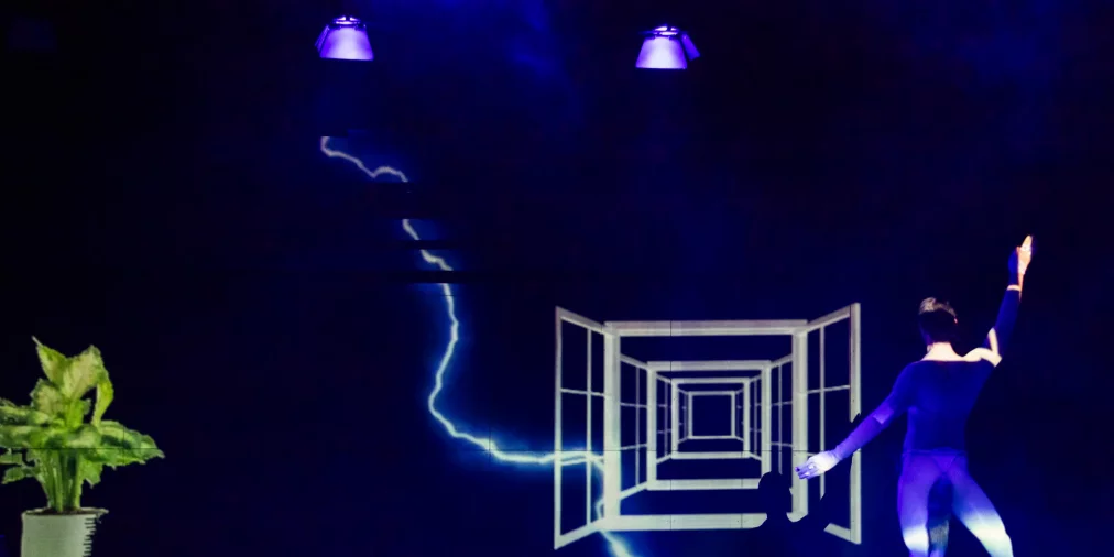 Eine Person steht auf einer dunklen Bühne vor einer großflächigen Projektion, die ein perspektivisch gestaffeltes Fenster- oder Tunnelmotiv zeigt. Neben der Projektion verläuft eine leuchtende, blitzartige Linie. Links am Bühnenrand steht eine grüne Zimmerpflanze in einem Topf. Die Person trägt enganliegende Kleidung und hebt einen Arm nach oben, während der andere Arm seitlich ausgestreckt ist. Violettes und blaues Bühnenlicht beleuchtet die Szene.