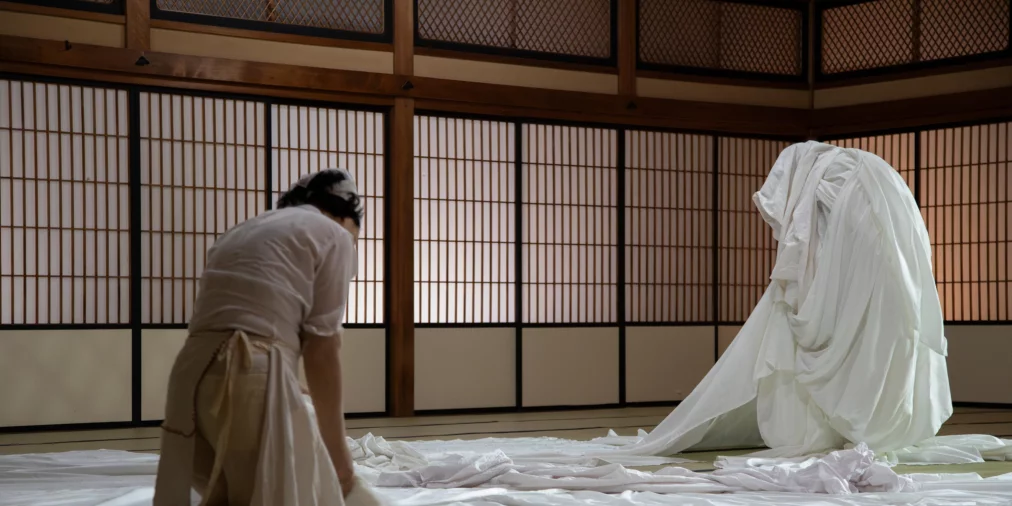 Zwei Personen arbeiten auf einem Boden voller weißer Stoffbahnen. Eine Person kniet und faltet Textilien, während eine zweite Person vollständig in weiße Stoffe gehüllt aufrecht sitzt. Traditionell wirkender Raum mit Holz und Schiebetüren im Hintergrund.