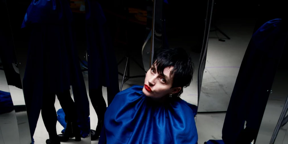 Eine Person mit kurzem dunklem Haar steht zwischen Spiegelpaneelen und blickt nach oben zur Kamera. Sie trägt ein weites, dunkelblaues Oberteil mit sehr langen Ärmeln, die auf dem Boden liegen, schwarze Beinkleidung und schwarze Stiefeletten. Die Spiegel vervielfachen Körper und Raum.