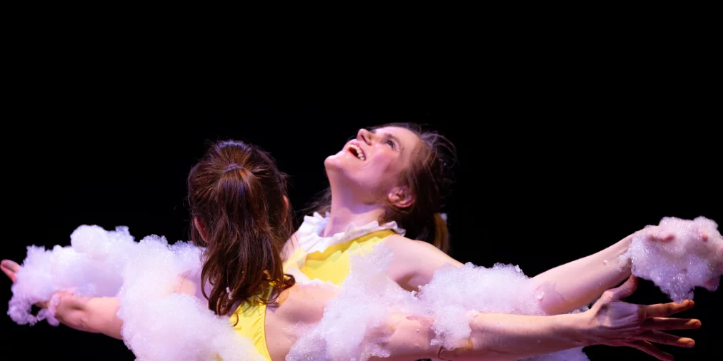Two performers stand back to back with their arms extended. Both wear yellow tops and are partially covered in white foam spread across shoulders, arms, and back. One person tilts their head backward and smiles while stage lighting highlights the scene.