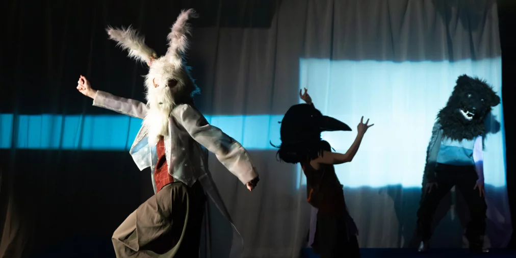 Three people wearing animal masks move across several platforms. One wears a white mask with long ears, another a black bird mask, and another a dark animal mask. Cool blue light shines through a curtain in the background.