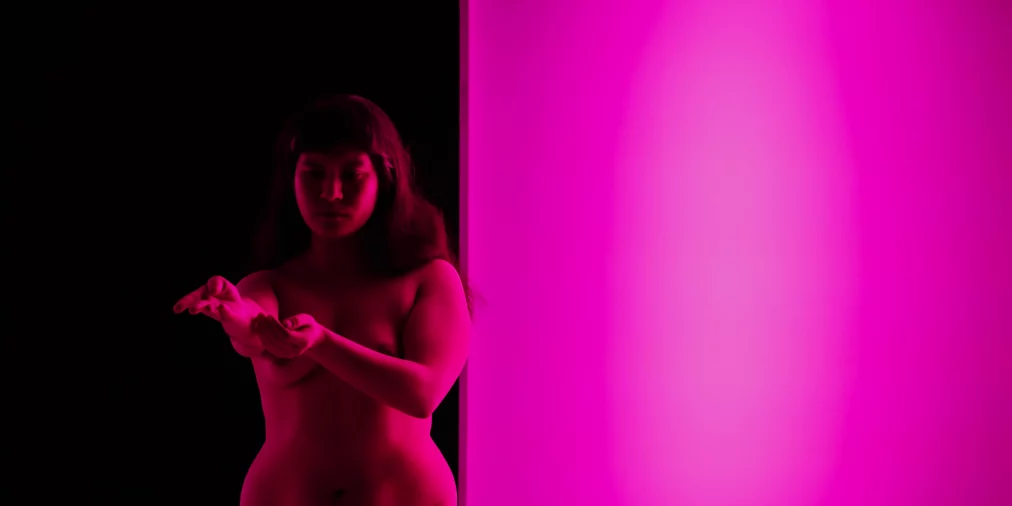 One person stands next to a glowing pink wall surface in a dark stage space. The body is illuminated by pink light while the background remains black. The person holds one hand in front of them and looks at it.