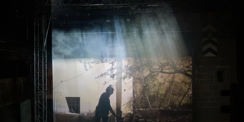 Eine große Videoprojektion zeigt die Silhouette einer Person, die sich in einem Hof oder Garten bewegt. Lichtstrahlen fallen von oben durch Bühnennebel und erzeugen sichtbare Lichtbündel über der Projektion.
