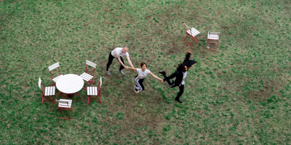 Three persons move across a grassy field while holding hands. The scene is photographed from above. Several white garden chairs and a round table with red frames stand nearby.