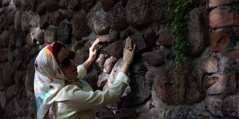 A person stands sideways in front of a rough stone wall, placing both hands flat against its uneven surface. They wear a light-colored outfit, a patterned headscarf, and sunglasses. Small green plants grow between the stones, while light and shadow structure the scene.
