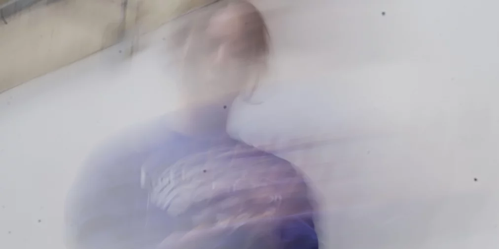 Eine Person ist durch starke Bewegungsunschärfe kaum erkennbar und scheint sich schnell vor einer hellen Wand zu bewegen. Die Konturen des Körpers lösen sich in weiche, verschwommene Formen auf.