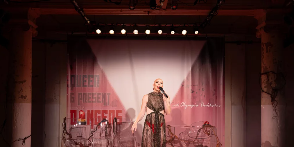 Eine Person steht auf einer roten Bühne und spricht in ein Mikrofon. Sie trägt ein langes, transparentes Kleid und rote Schuhe. Hinter ihr stehen transparente Stühle und ein Tisch mit dekorativen, wurzelartigen Elementen. Im Hintergrund ist ein Schriftzug sichtbar.