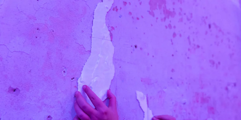 Zwei Hände berühren die Oberfläche einer Wand unter violettem Licht. Teile der Wand sind aufgebrochen und geben strukturierte, reliefartige Formen frei. Die Hände tasten vorsichtig über die unterschiedlichen Oberflächen.
