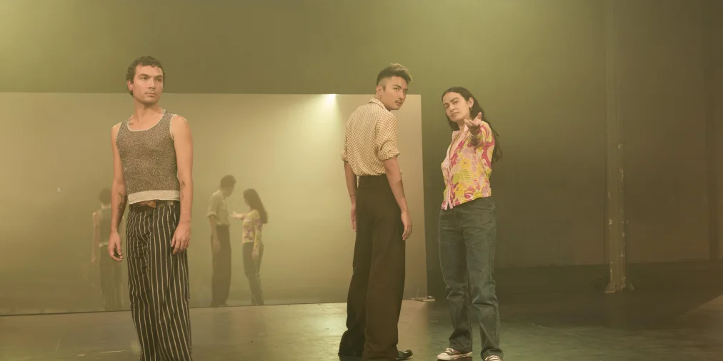 Three people stand on a hazy stage lit in greenish tones. On the left, a person with curly hair, a glittery tank top and striped trousers looks to the side. On the right, a person in a checked shirt glances over their shoulder, next to a person in a floral blouse and jeans who extends one hand forward. A large wall behind them mirrors the group.