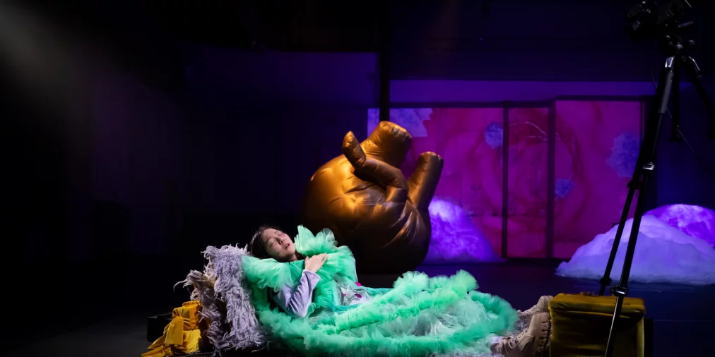 Performer in a green tulle costume lies on stage surrounded by props, with a large inflatable object and colorful illuminated stage set in the background.