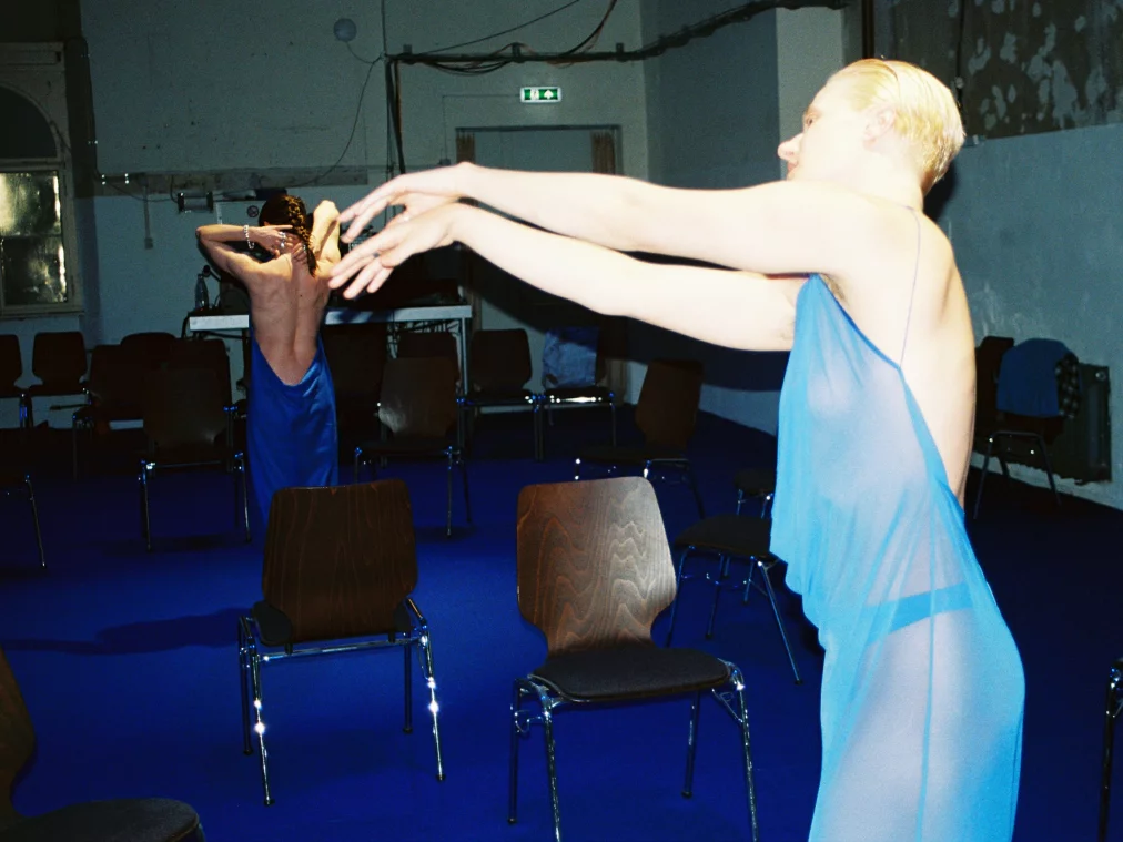 Two people in blue dresses are standing in a room with lots of empty chairs and a blue carpet.