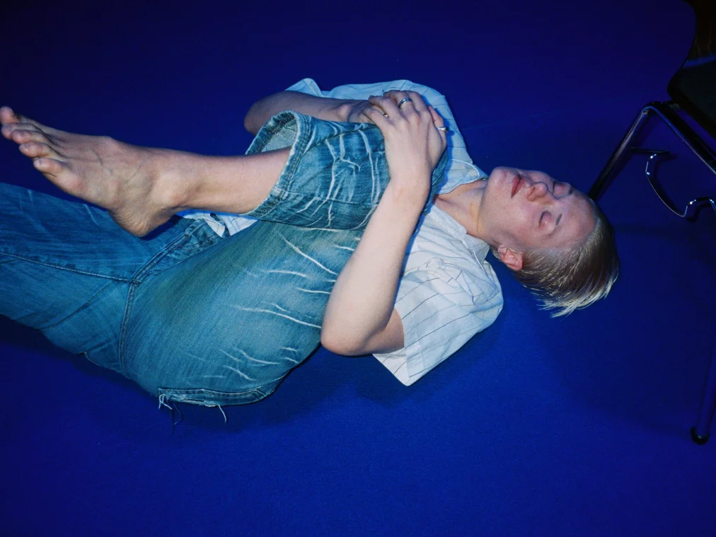 A person is lying on a blue carpet. They pull one leg to their chest, their eyes are closed.