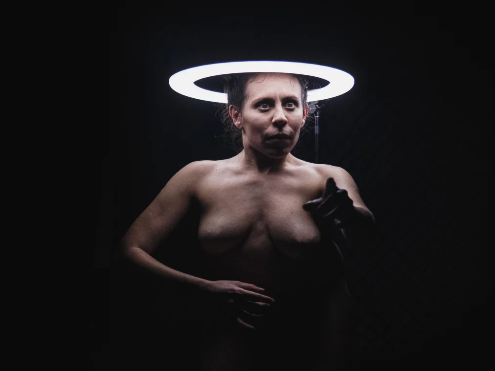 A nude performer stands against a dark background beneath a glowing ring-shaped light positioned above her head, resembling a halo. She looks intently forward and points toward the camera.