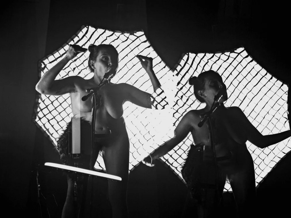  A single performer with a bare upper body appears twice in the image, standing at two microphones in front of illuminated mesh-pattern backdrops. She wears a striking stage accessory at the hip and gestures expressively with her arms. The photo is in black and white.