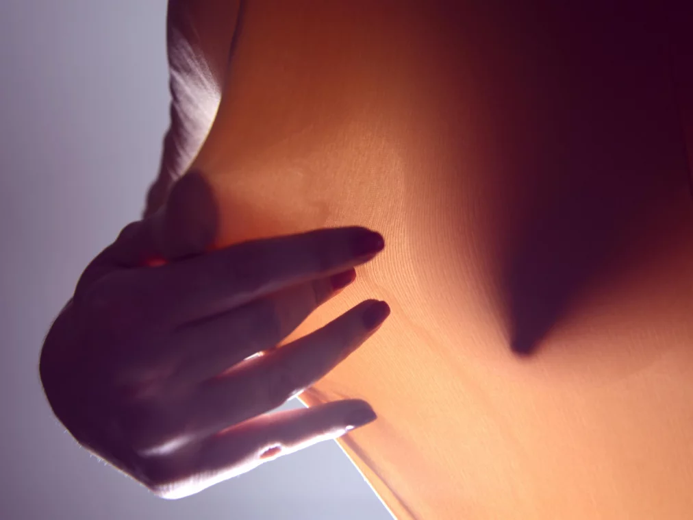 Eine Nahaufnahme zeigt eine Hand, die ein Stück transparenten, orangefarbenen Stoff sanft berührt. Das Licht von hinten lässt die feine Struktur des Stoffes erkennen und wirft den Schatten der Hand deutlich auf die Oberfläche.