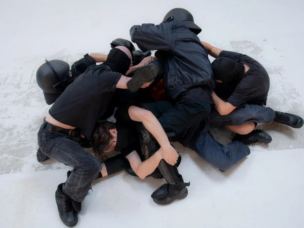 Mehrere Personen liegen auf einem weißen Boden. Einige tragen schwarze Kleidung mit Helmen. Arme und Beine sind ineinander verhakt, der Boden ist leer bis auf die Personen.