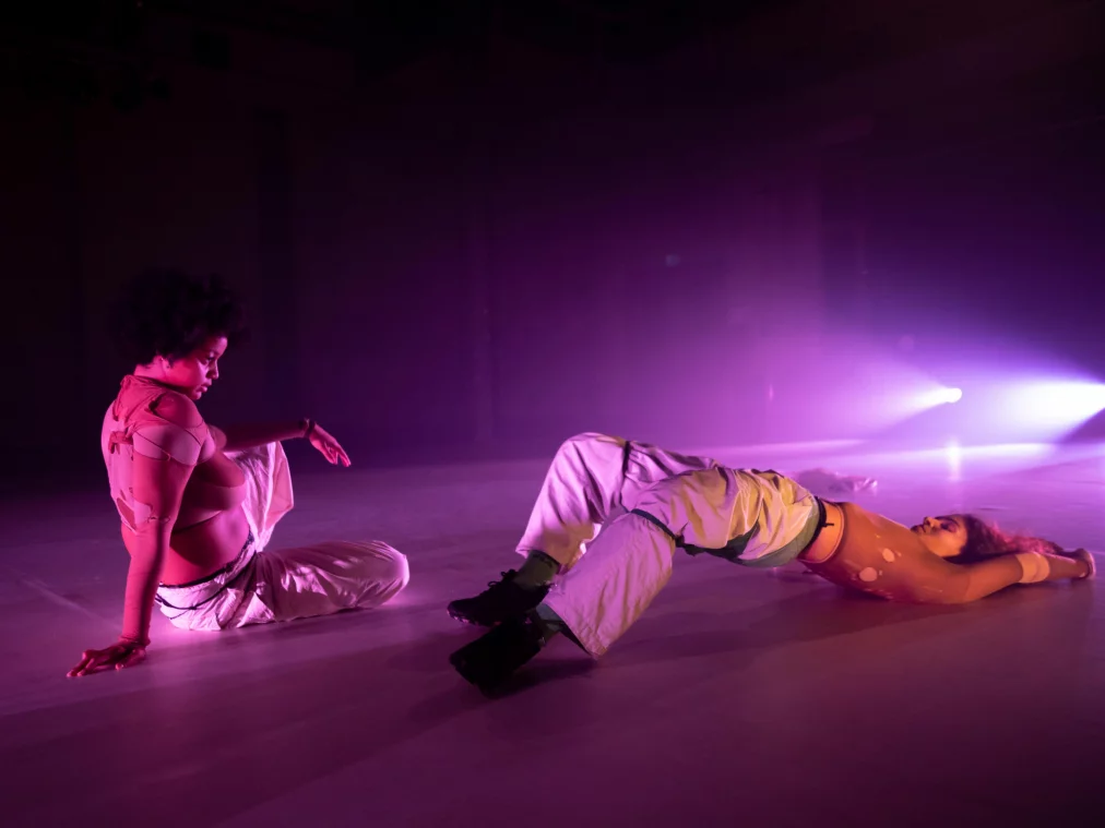 Zwei Performer*innen liegen und sitzen auf einer Bühne, in violettem Licht. Eine Person liegt auf dem Rücken, die andere sitzt nah daneben und stützt sich mit einer Hand ab.