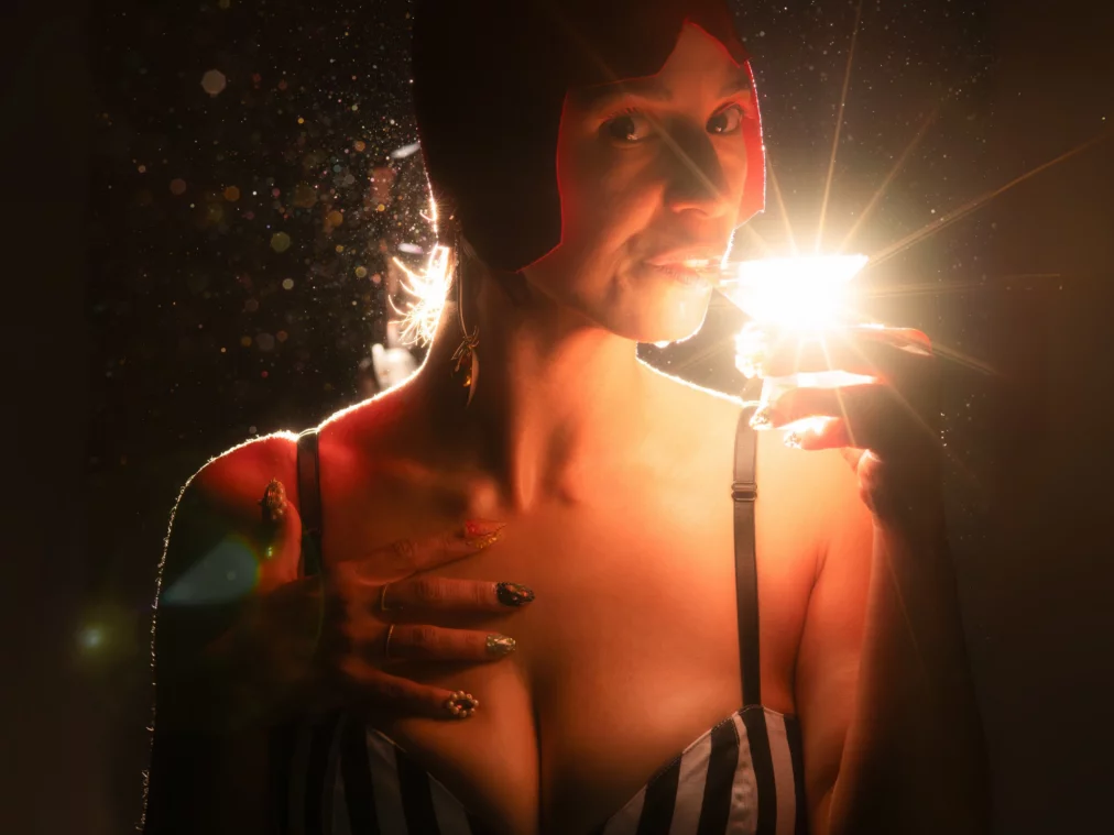 Person in a striped top and red cap holds a cocktail glass illuminated by bright light.