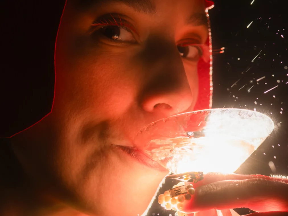 Close-up of a person wearing a red cap, drinking from a cocktail glass, with sparks in the background.