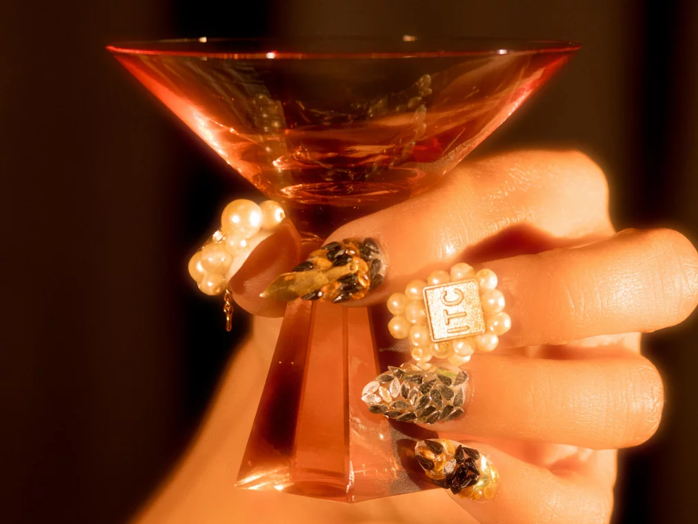 Nahaufnahme einer Hand, die ein rotes Cocktailglas hält, mit langen, verzierten Fingernägeln.