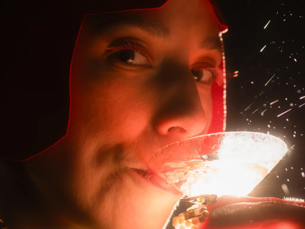 Nahaufnahme einer Person mit roter Kappe, die aus einem Cocktailglas trinkt, Funken im Hintergrund.