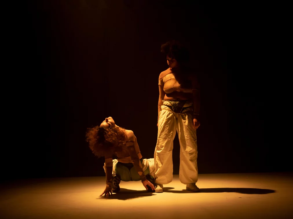 Two people in skin-colored tops with round cutouts and loose trousers on a dimly lit stage. One person kneels with their upper body leaning back, while the other stands upright in front of them, looking down.