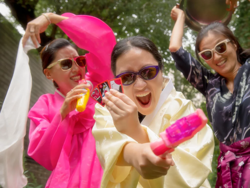 Three people in colorful hanbok, traditional Korean clothing with wide sleeves and bright colors, and sunglasses pose outdoors, one pointing playing cards and a water gun towards the camera.