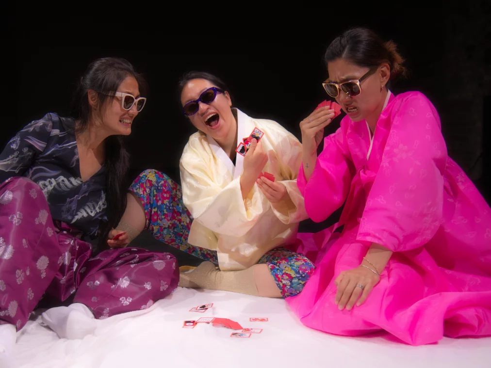 Three people in colorful hanbok, traditional Korean clothing with wide sleeves and bright colors, and sunglasses sit laughing on a white cloth, holding playing cards.