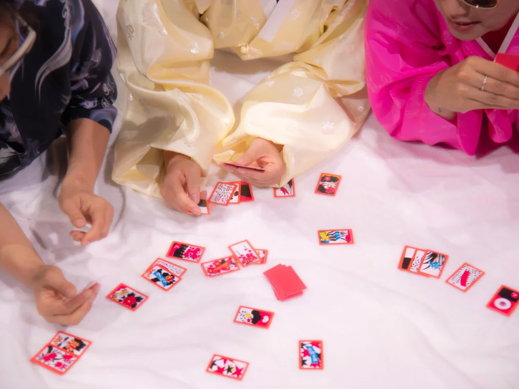Close-up of the hands of three people in colorful hanbok, traditional Korean clothing with wide sleeves and bright colors, sorting playing cards on a white cloth.