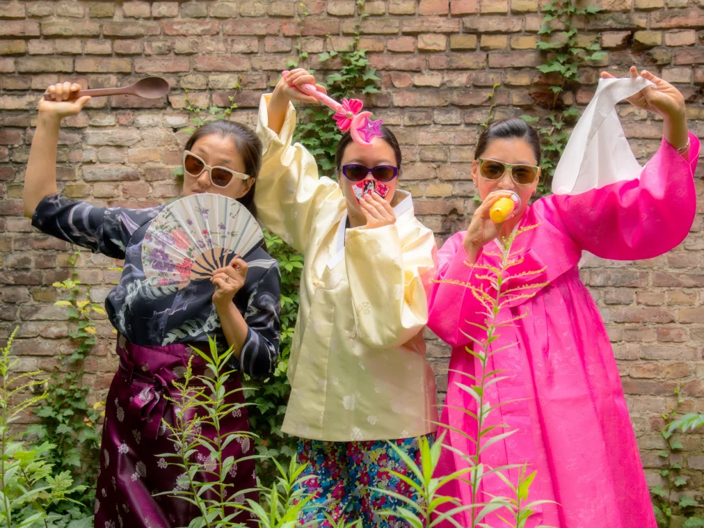 Three people in colorful hanbok, traditional Korean clothing with wide sleeves and bright colors, and sunglasses pose in front of a brick wall, each holding different props.