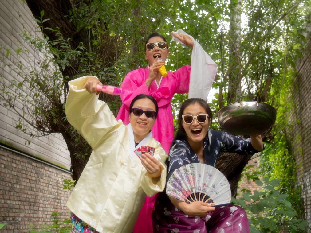 Three people in colorful hanbok, traditional Korean clothing with wide sleeves and bright colors, and sunglasses pose outdoors, one holding a cooking pot, another holding playing cards.