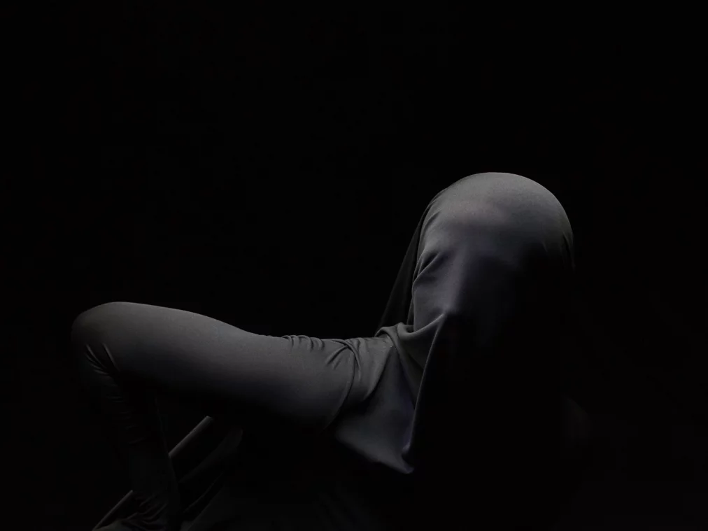 A Person is entirely covered by a black cloth that clings closely to the body, revealing its contours. They are positioned in a bent posture against a black background.