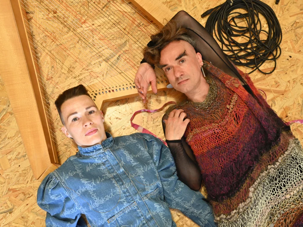 Two people lie on a wooden floor next to a large wooden string structure. The person on the left wears a denim top, the person on the right a multicolored knitted top with sheer sleeves. A coiled cable lies beside them.