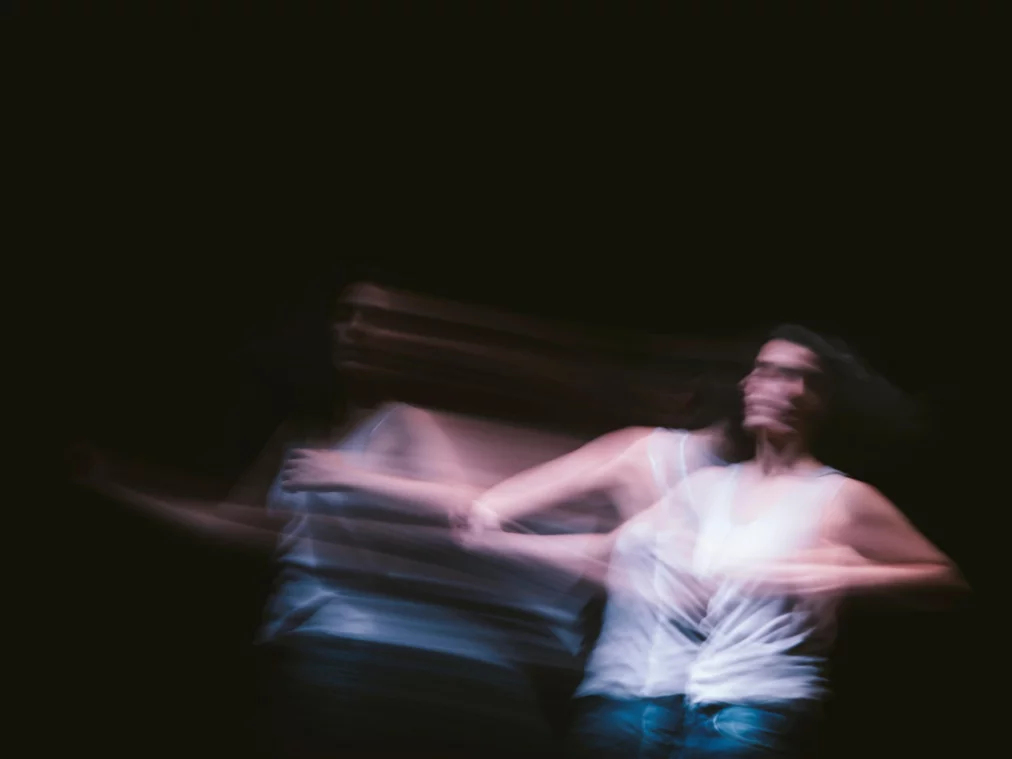 Verschwommene Aufnahme einer Person in Bewegung vor schwarzem Hintergrund. Die Person trägt ein weißes Oberteil und blaue Hose, die Bewegungen lassen mehrere überlagerte Silhouetten entstehen.