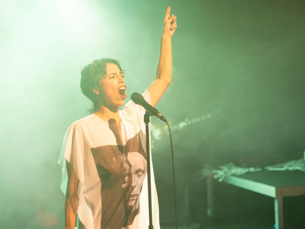 A person with short dark hair stands in stage fog at a microphone stand. They wear a white T-shirt with a printed portrait and raise their right arm with an extended finger upwards.