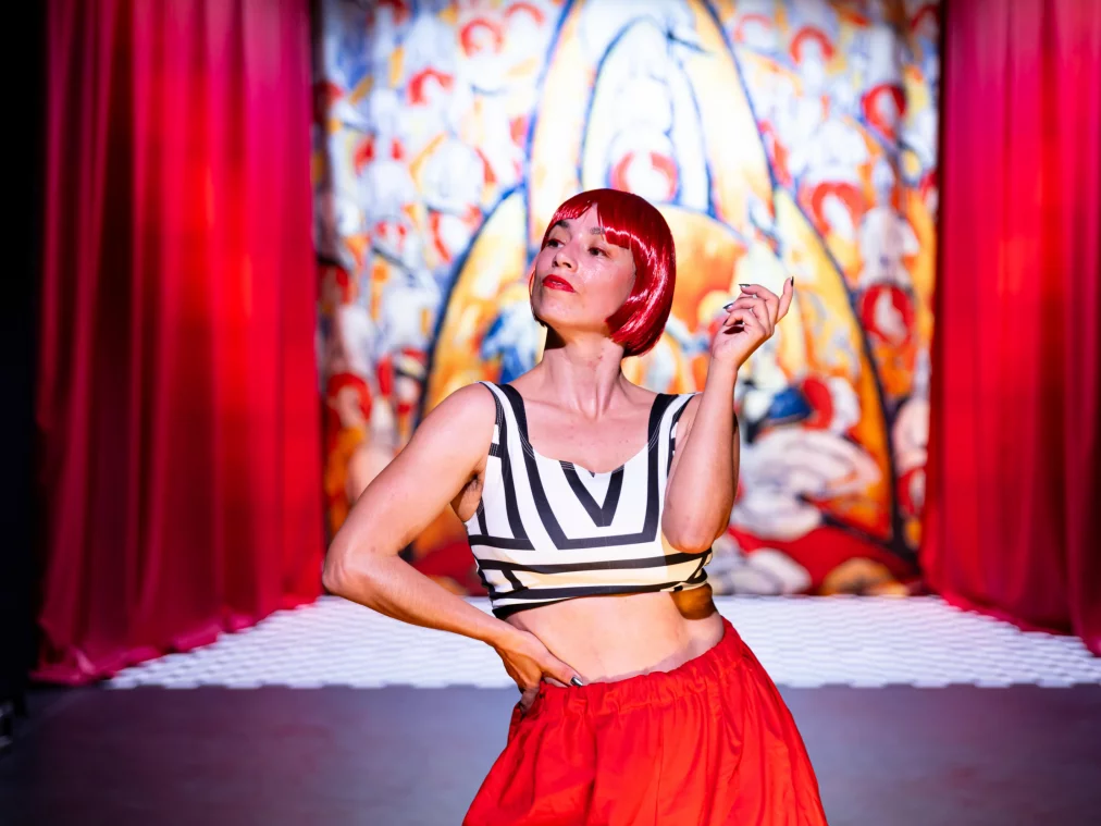 A person with a red wig stands in front of a colorful stage backdrop between red curtains. They wear a black-and-white striped top and a red skirt, with one hand on their hip and the other slightly raised.