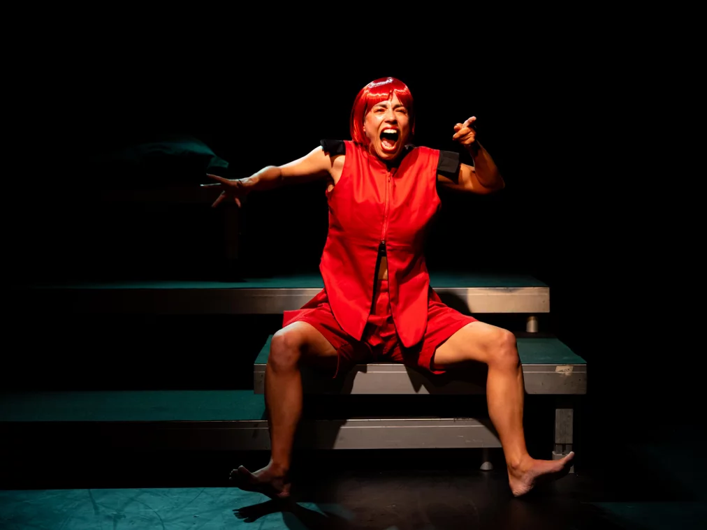 A person with a red wig sits with legs apart on a stage platform. They wear a red outfit without shoes and shout with their mouth wide open, arms stretched forward.