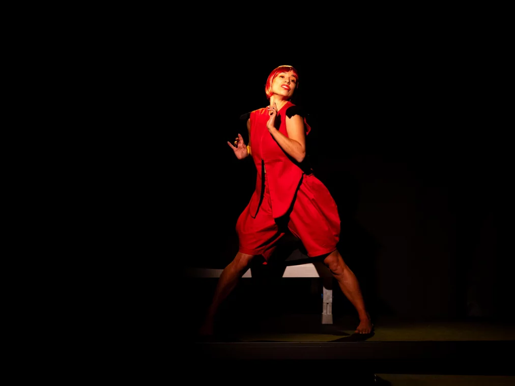 A person with a red wig stands barefoot on a dark stage. They wear a red outfit and pose with bent arms, head slightly tilted to the side and a smile on their face.