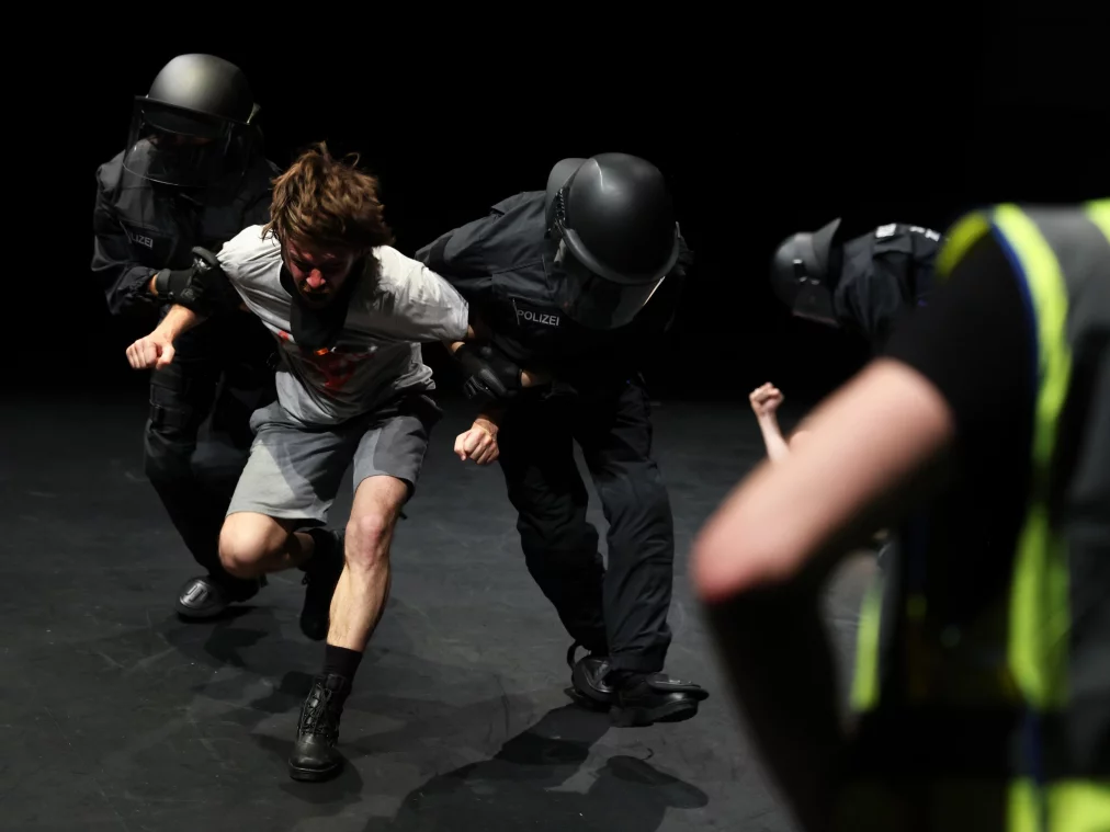 Two people in black protective gear and helmets hold a person in a gray T-shirt and shorts by the arms, pulling them forward. Another person in a yellow safety vest stands in the foreground with their back to the camera.