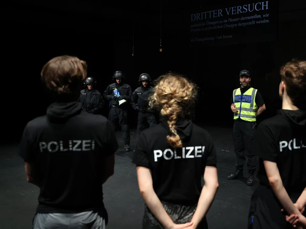 Several people stand on a stage. In the foreground, three people wear black T-shirts with “POLIZEI” printed on them. Behind them, four people in black protective gear with helmets stand. A person in a yellow safety vest speaks to them. Above the scene, projected text displays the heading “DRITTER VERSUCH.”