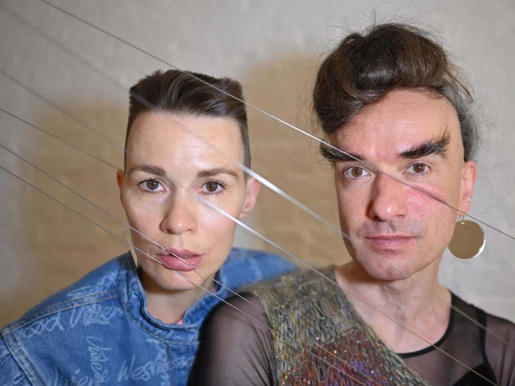 Nahaufnahme von zwei Personen hinter gespannten Saiten, die quer vor ihren Gesichtern verlaufen. Links trägt die Person ein Jeansoberteil, rechts die Person ein gestricktes Oberteil und einen runden Ohrring.