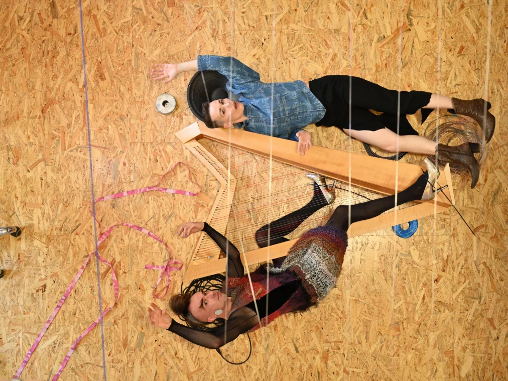 Zwei Personen liegen auf einem Holzfußboden zwischen Kabeln und einer großen Holzkonstruktion mit Saiten. Eine Person trägt ein blaues Jeansoberteil, die andere ein bunt gestricktes Oberteil mit transparenten Ärmeln.